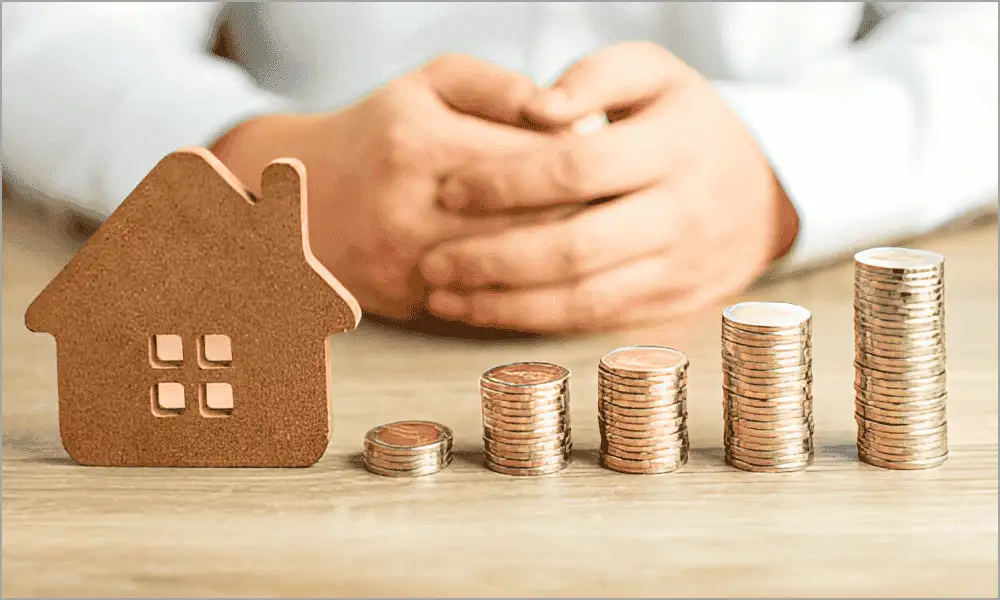A man sitting at a desk with a small house model and coins arranged in ascending order, symbolizing increasing interior real estate photography prices