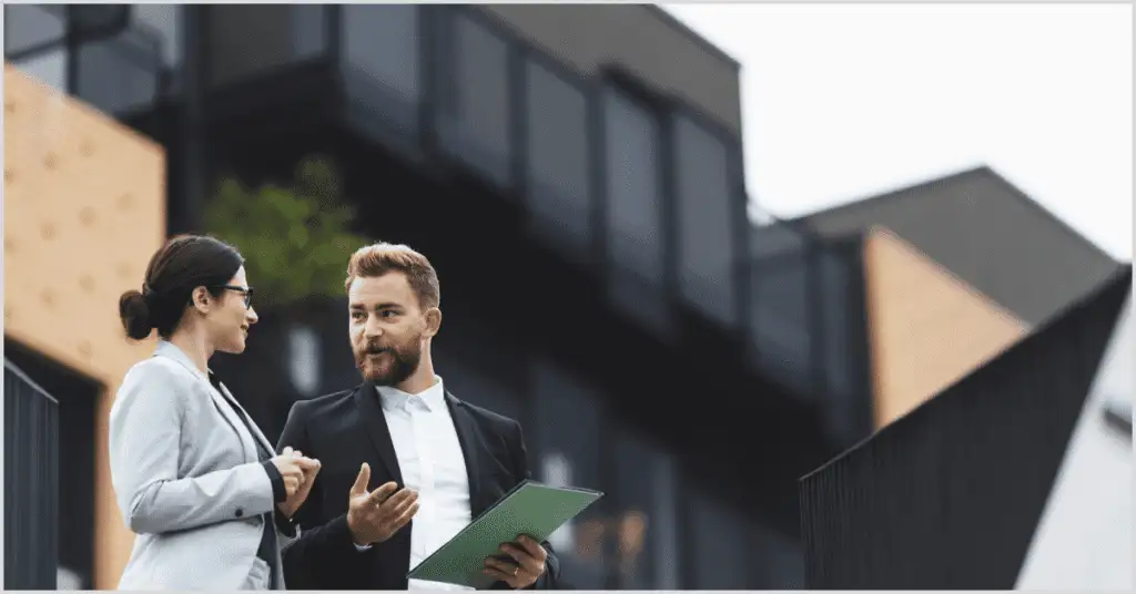 Two professionals in suits discuss a project with a green folder, standing in front of modern buildings, representing collaboration in real estate renderings.