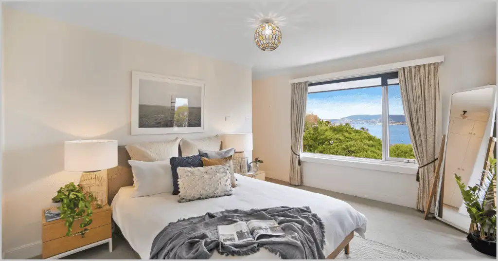 A bright, modern bedroom with a large window offering a stunning view of greenery and a serene water body. The room features a neatly made bed with white linens, colorful pillows, and a gray throw blanket, accompanied by a wooden nightstand with a lamp and potted plant.