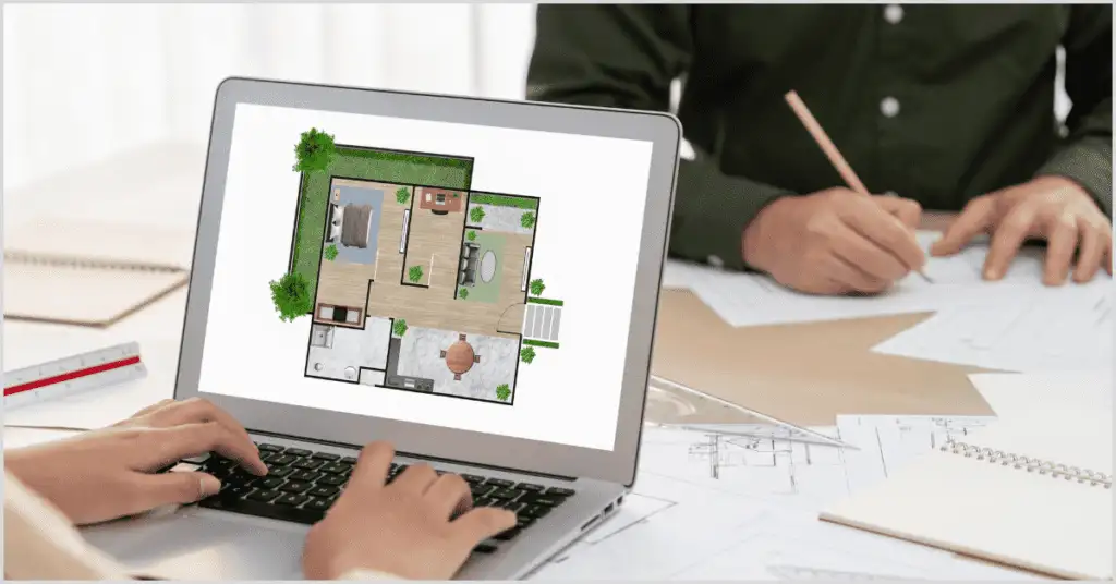 A laptop showing a 3D Real Estate Floor Plan Drawing of a furnished home, with hands typing and sketching on paper blueprints nearby.