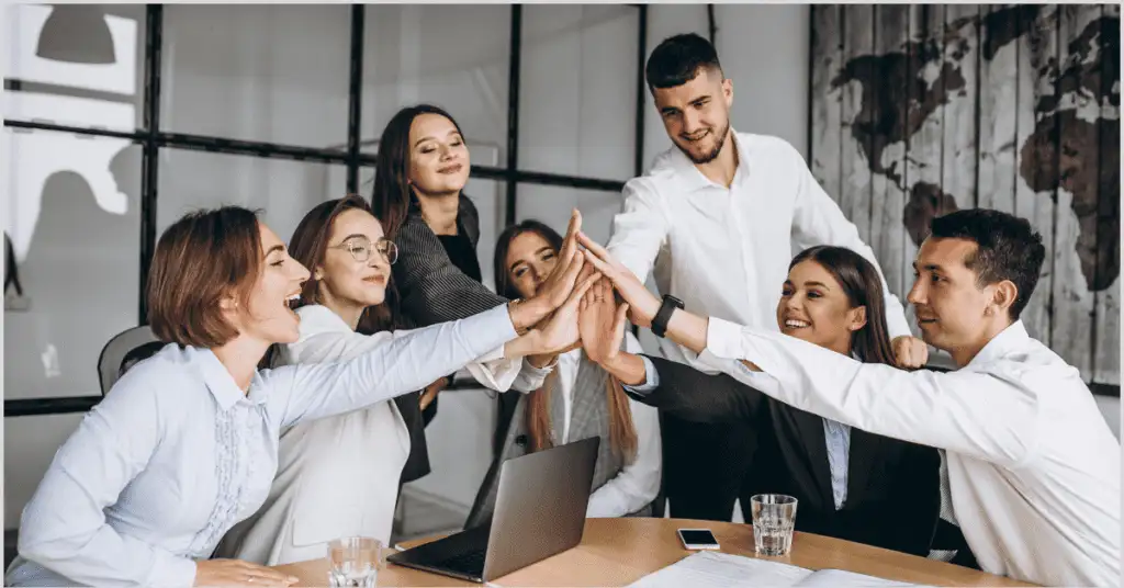 A group of professionals in a modern office giving high-fives around a conference table with laptops, documents, and water glasses, celebrating teamwork related to finding real estate buyers.