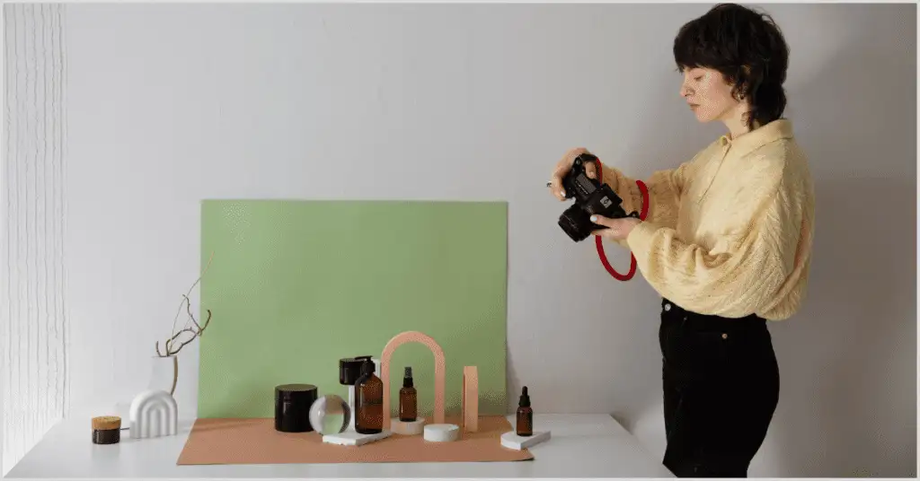 A person with a camera photographs a styled setup with skincare products, a white vase, and a green backdrop on a table.
