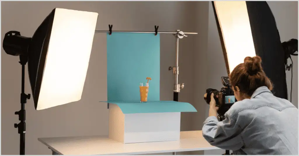 A person uses a camera to photograph a table setup with skincare products, a white vase, and a green backdrop for a branded shoot.