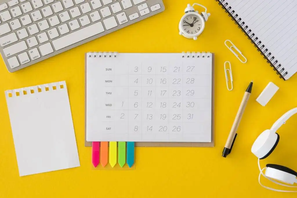 A desk setup with a calendar, keyboard, headphones, notebooks, a clock, and colorful sticky notes on a yellow background.