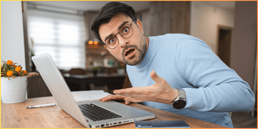 Image of a surprised man in a blue sweater working on a laptop at a desk with a plant and phone nearby.