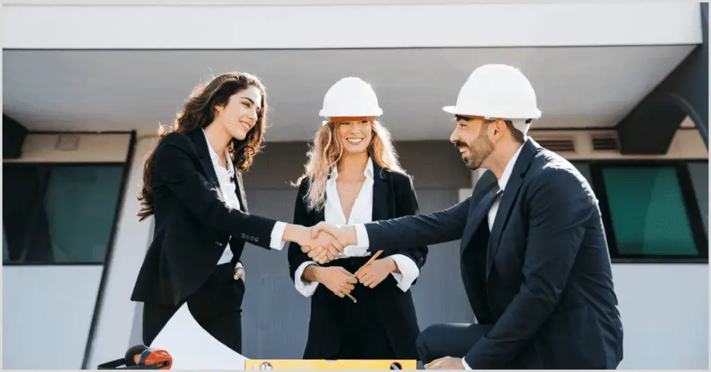Three professionals in hard hats shaking hands in front of a building, symbolizing partnership with home builders.