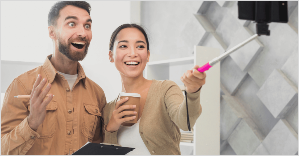 Two excited people recording a live video with a selfie stick, holding coffee and a clipboard, promoting open house ideas.