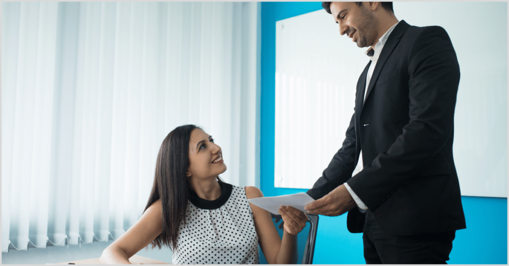 A man in a suit handing a document to a woman in a polka-dot dress in an office, representing a home buying timeline.