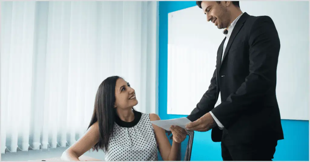 A man in a suit handing a document to a woman in a polka-dot dress in an office, representing a home buying timeline.