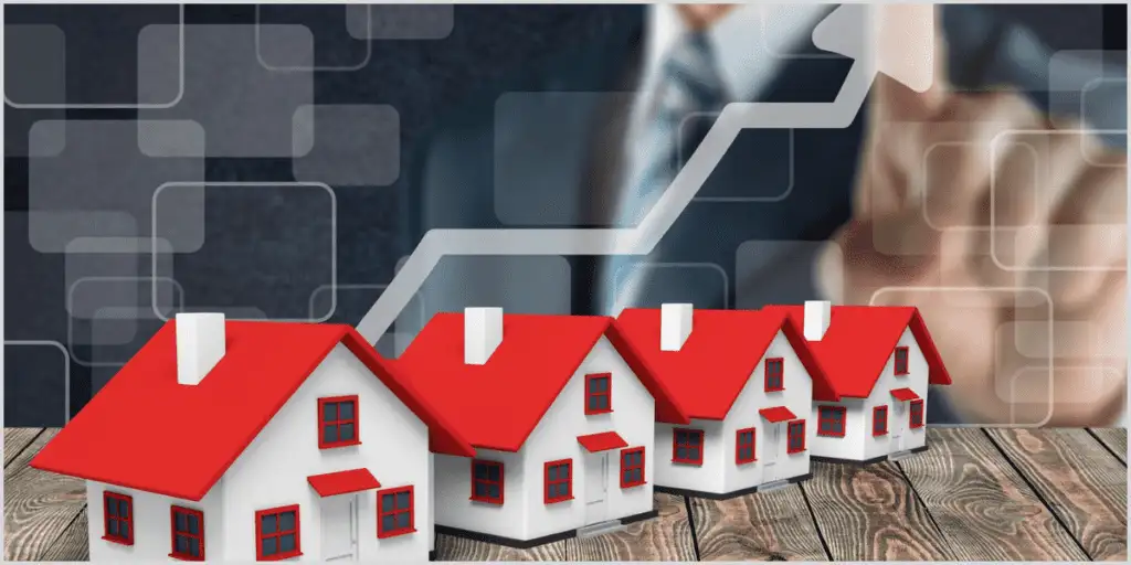 A row of small model houses with red roofs on a wooden surface, with a blurred background featuring an upward-trending graph and a businessperson's hands, symbolizing Real Estate Investing.