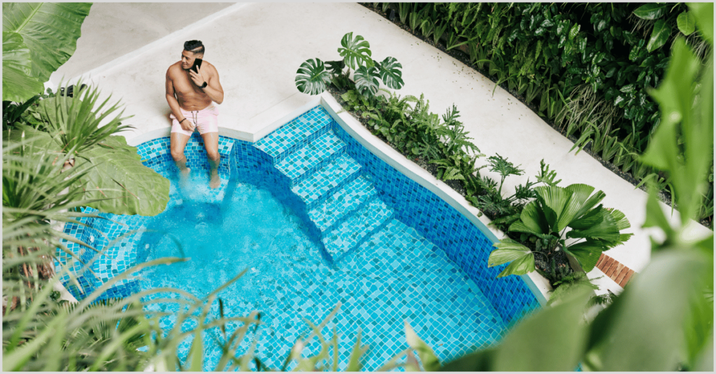 Man sitting by a pool while talking on the phone surrounded by lush greenery.