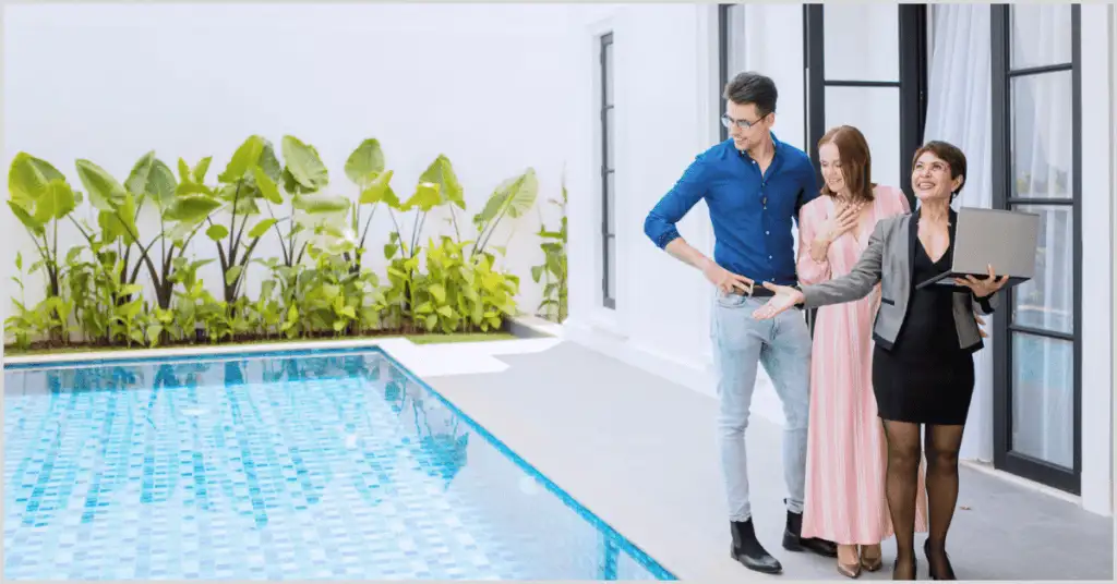 Three people discussing by a pool with low-maintenance greenery, one holding a laptop, near a modern house with large windows.