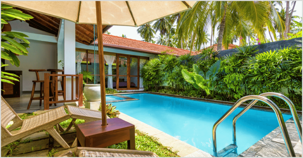 A serene pool area surrounded by low-maintenance greenery, featuring a clear blue pool, wooden lounge chairs, and a shaded patio with palm trees in the background.