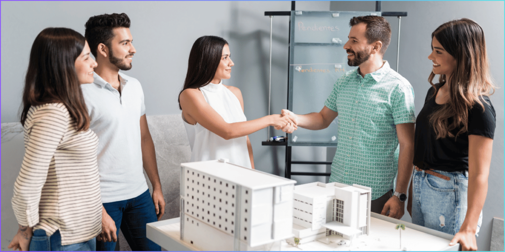 Diverse group of four professionals in a modern office shaking hands over a white architectural model of a multi-story building, with a glass board labeled "Pendientes" (pending) in the background, surrounded by a blue border.