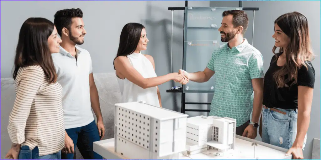 Diverse group of four professionals in a modern office shaking hands over a white architectural model of a multi-story building, with a glass board labeled "Pendientes" (pending) in the background, surrounded by a blue border.