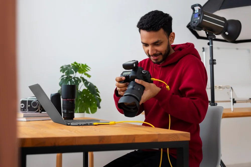 A young man in a red hoodie sitting at a desk, smiling as he connects his DSLR camera to a laptop with a yellow cable in a cozy home studio setup.