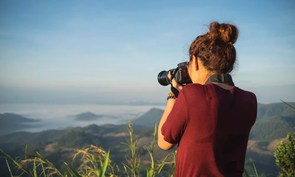 A woman with her hair in a bun, wearing a maroon shirt, photographing a misty mountain landscape under a clear sky.