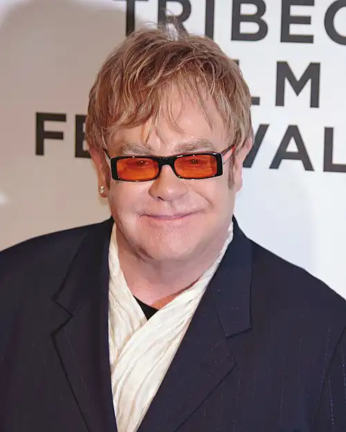 Elton John at Tribeca Film Festival, signature orange-tinted sunglasses, blonde hair and black suit, smiling – Who Is The Most Photographed Person In The World