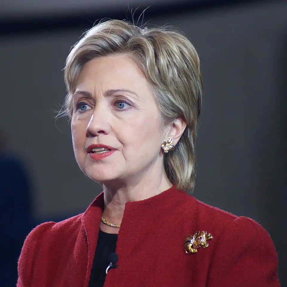 Hillary Clinton speaking at event, short blonde hair, red jacket and gold brooch, serious expression