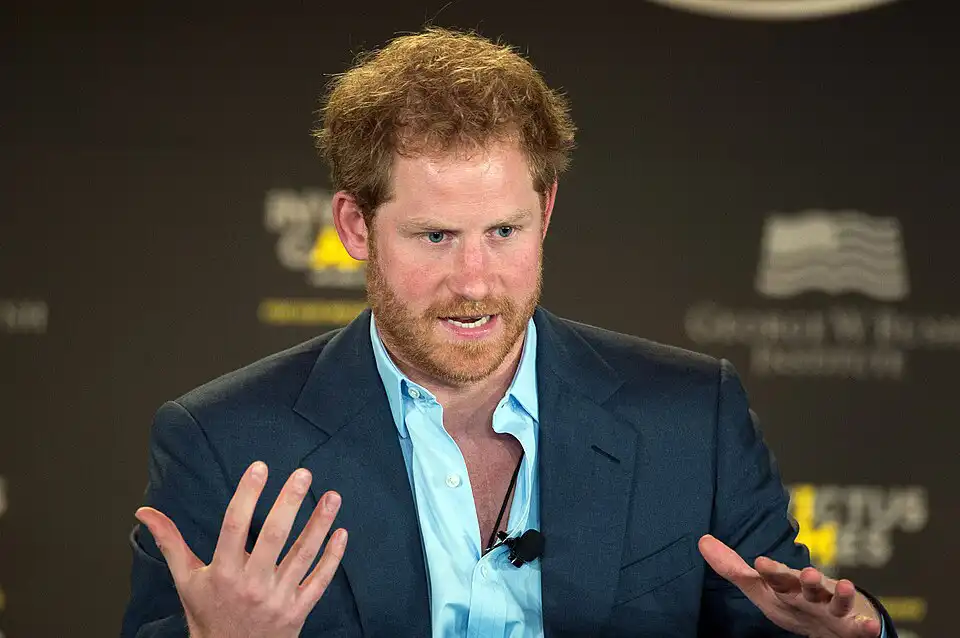 Prince Harry speaking passionately at a panel event, gesturing with hands, red hair and beard in navy suit – Who Is The Most Photographed Person In The World