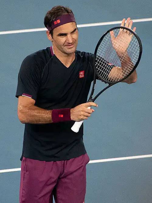 Roger Federer waving on tennis court in black UniQlo shirt and maroon shorts, holding racket, smiling – Who Is The Most Photographed Person In The World