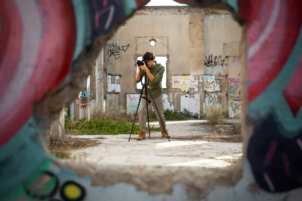A photographer on a tripod capturing images in an abandoned building covered in colorful graffiti, viewed through a broken wall with vibrant street art framing the scene.