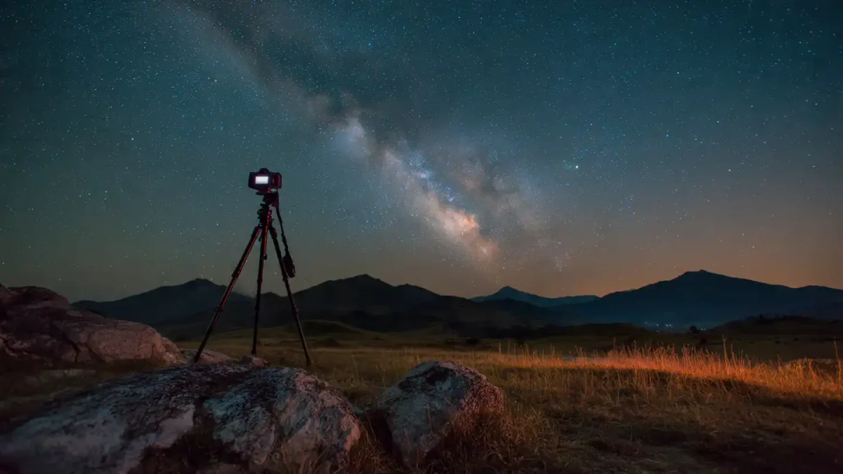 DSLR camera mounted on a tripod in a rocky highland landscape at night, rear LCD screen brightly lit showing a sharp, detailed preview of the glowing Milky Way arching across a deep starry sky, subtle twilight glow on distant mountain ridges, camera set for manual focus with live view active.